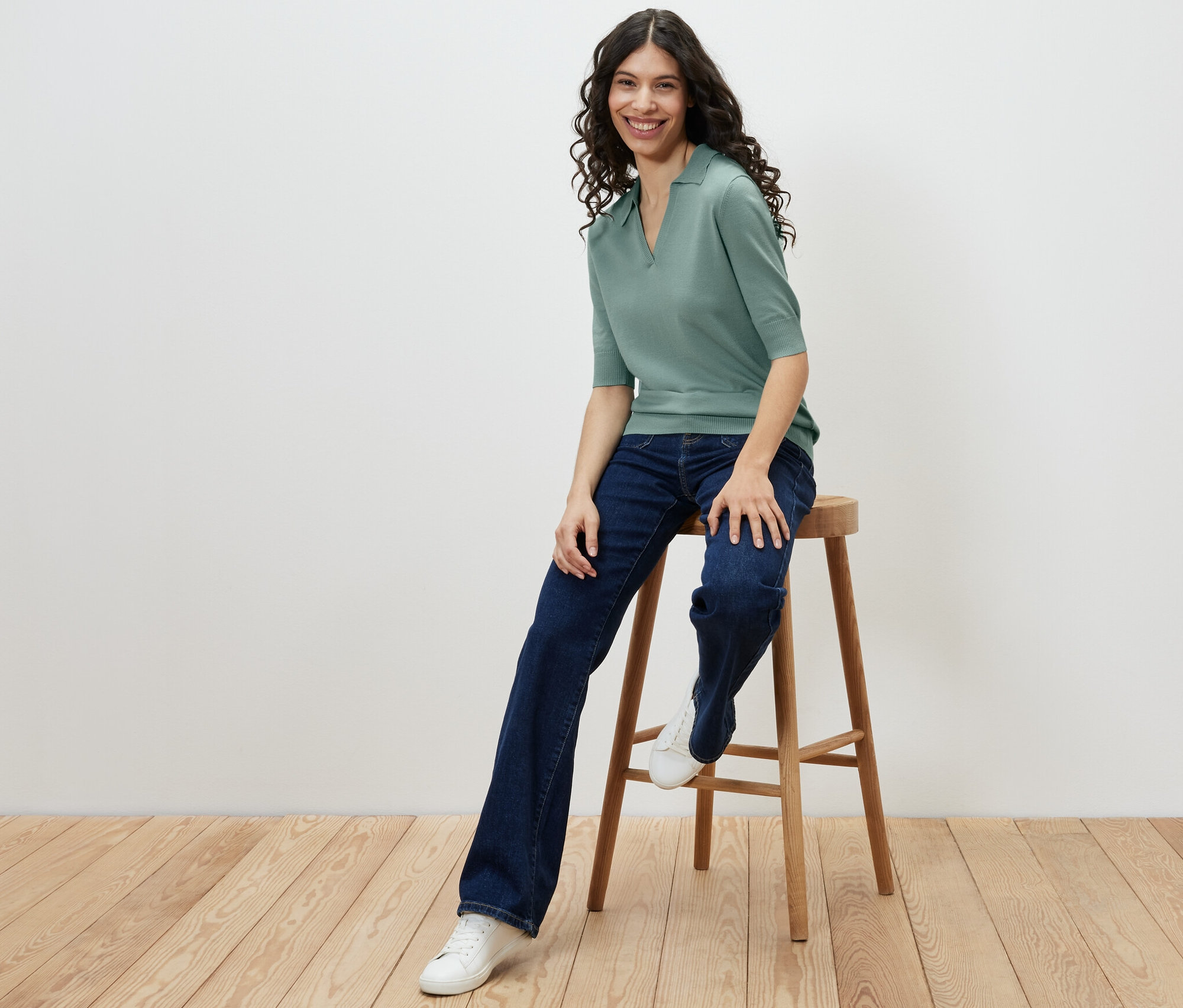 Nő ül egy széken, finom kötésű pólót, farmert és tornacipőt visel.