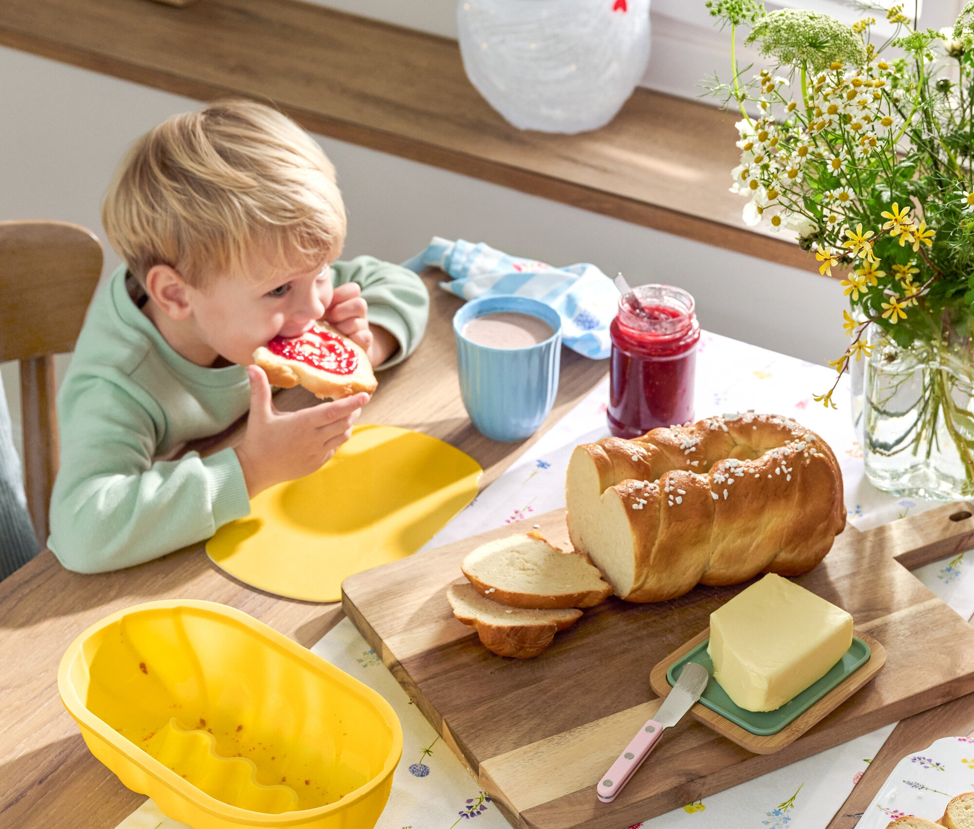 Szőke fiú lekváros kenyeret eszik az asztalnál kaláccsal, vajjal, lekvárral, edényekkel és virágokkal.