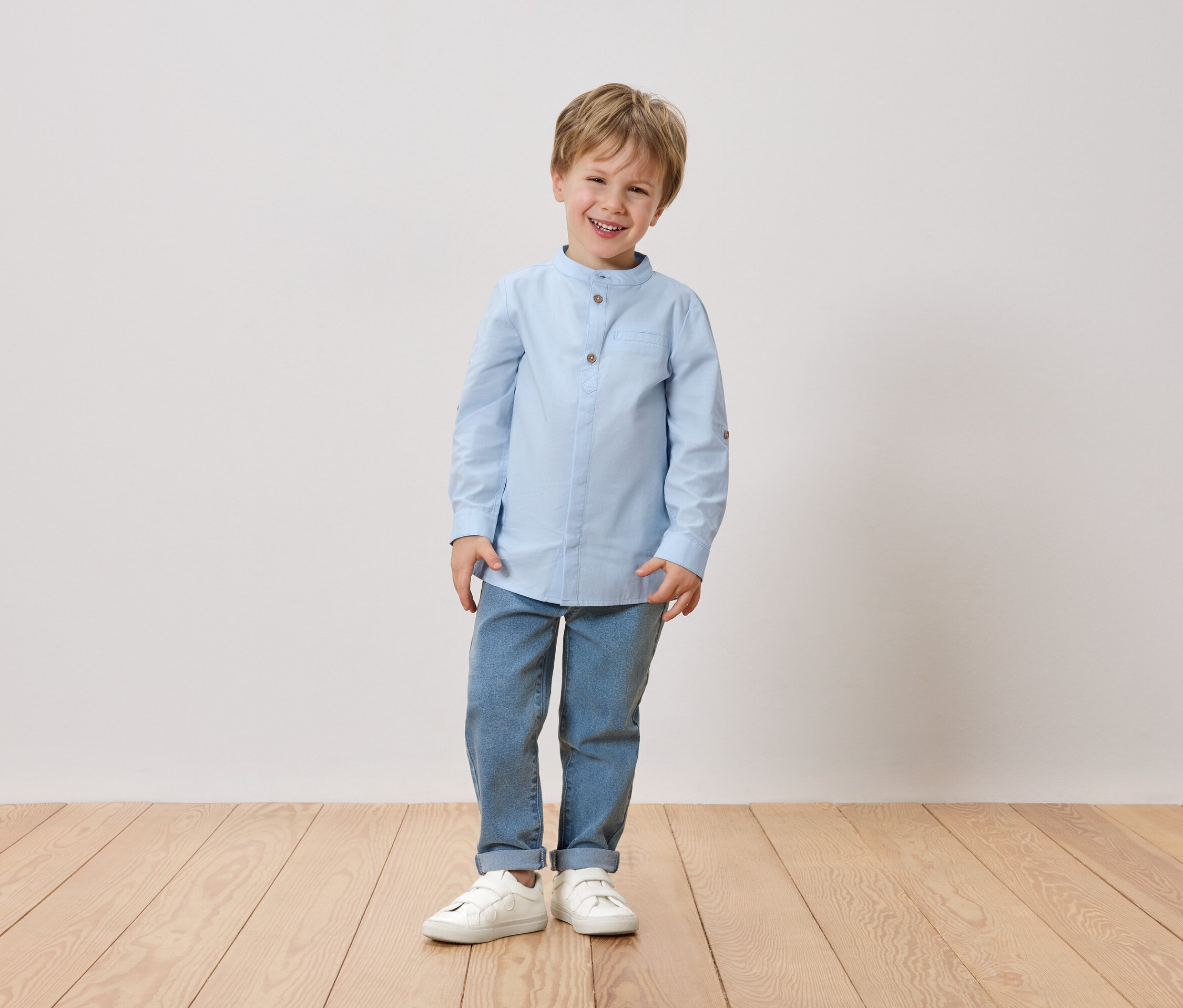 Egy mosolygó, szőke hajú fiú áll egy fehér fal előtt. Világoskék inget, farmert és fehér cipőt visel.