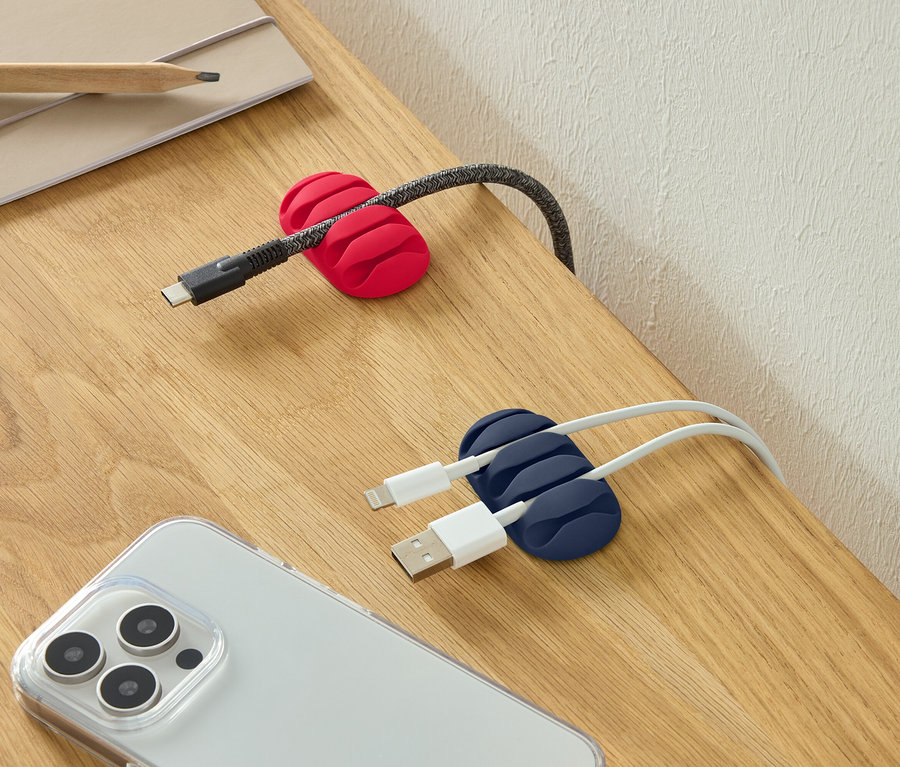 Egy USB-C kábel egy piros kábelrögzítőben, két USB-kábel egy kék kábelrögzítőben és egy okostelefon egy asztalon.