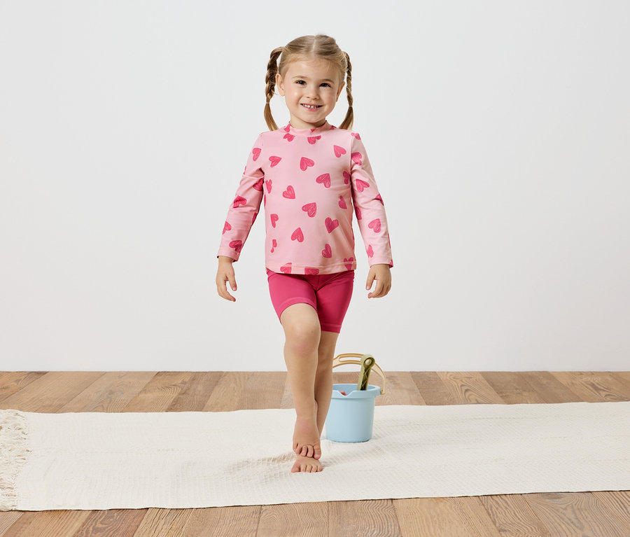 Fonott hajú kislány rózsaszín, szívecskés felsőben és rövidnadrágban egyensúlyoz egy fehér szőnyegen, egy világoskék vödör mellett.
