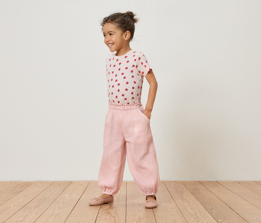 Egy kislány áll a fapadlón rózsaszín epres pólóban és rózsaszín nadrágban.