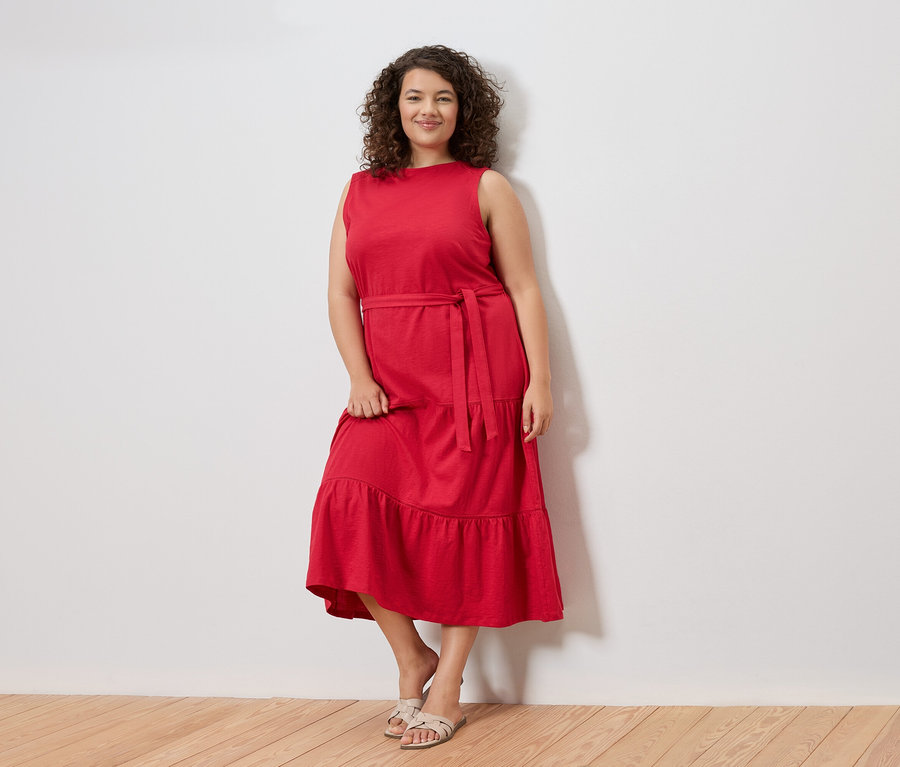 Egy göndör hajú nő áll egy fehér fal előtt, piros, ujjatlan ruhát visel övvel és szandállal.