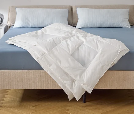 Egy kék lepedővel és két párnával ellátott ágyon egy fehér paplan fekszik. Az éjjeliszekrényen egy ventilátor áll.