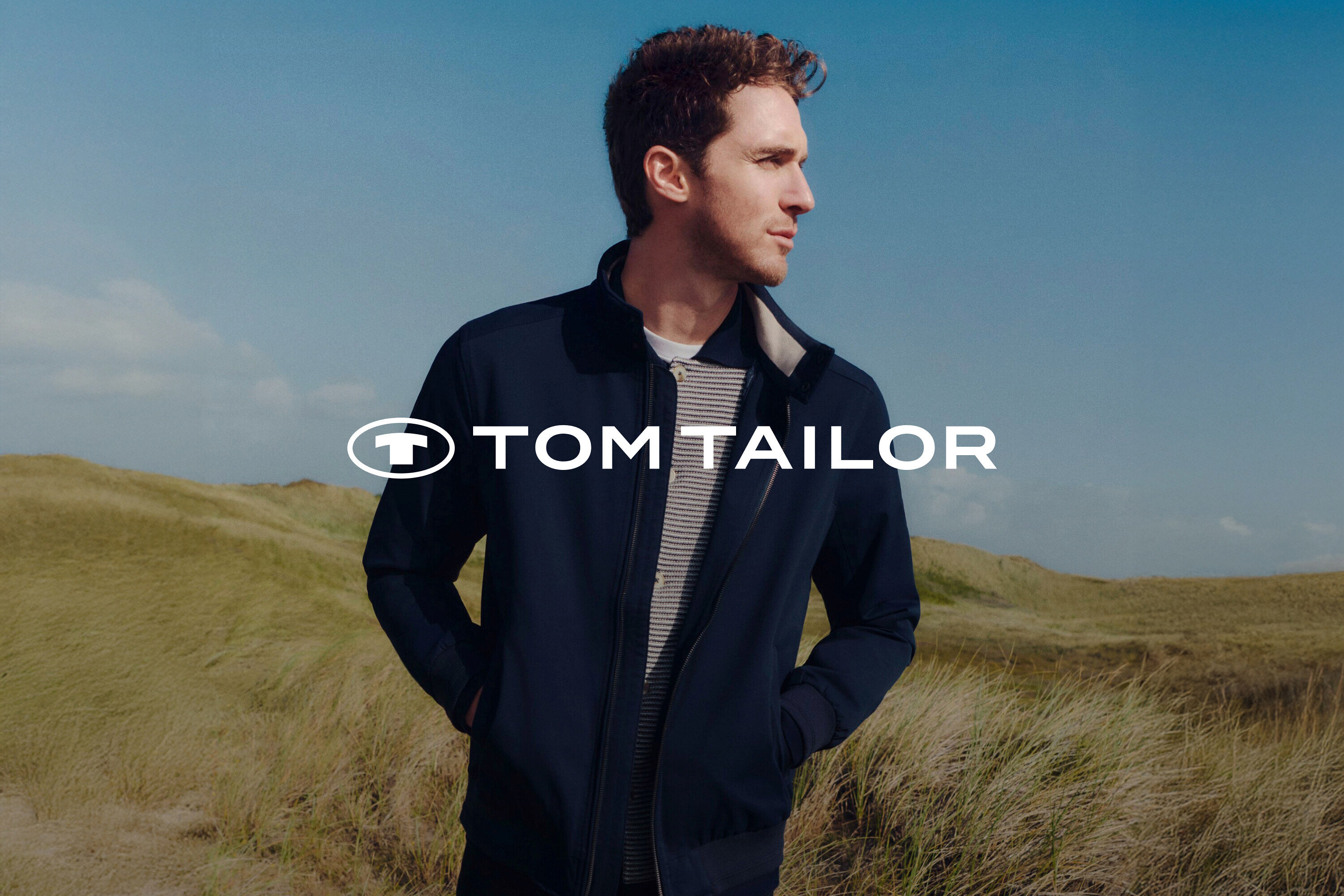 Férfi sötétkék kabátban és csíkos ingben, füves táj és kék ég előtt.