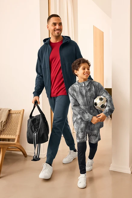 Férfi sporttáskával és gyerek labdával, mindketten sportruházatot viselnek. A férfi Sweatjacke-t és Sweathose-t visel. A gyerek Kinder-Windbreaker-Jacke-t visel.