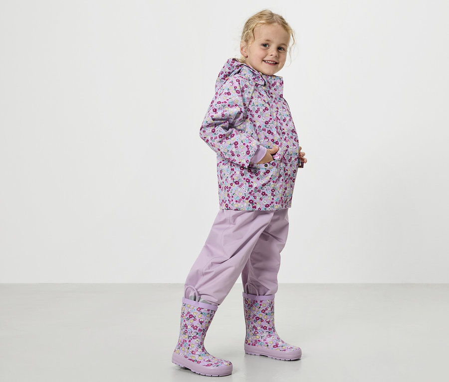Egy szőke kislány virágmintás lila esőkabátot, lila esőnadrágot és hozzáillő gumicsizmát visel.