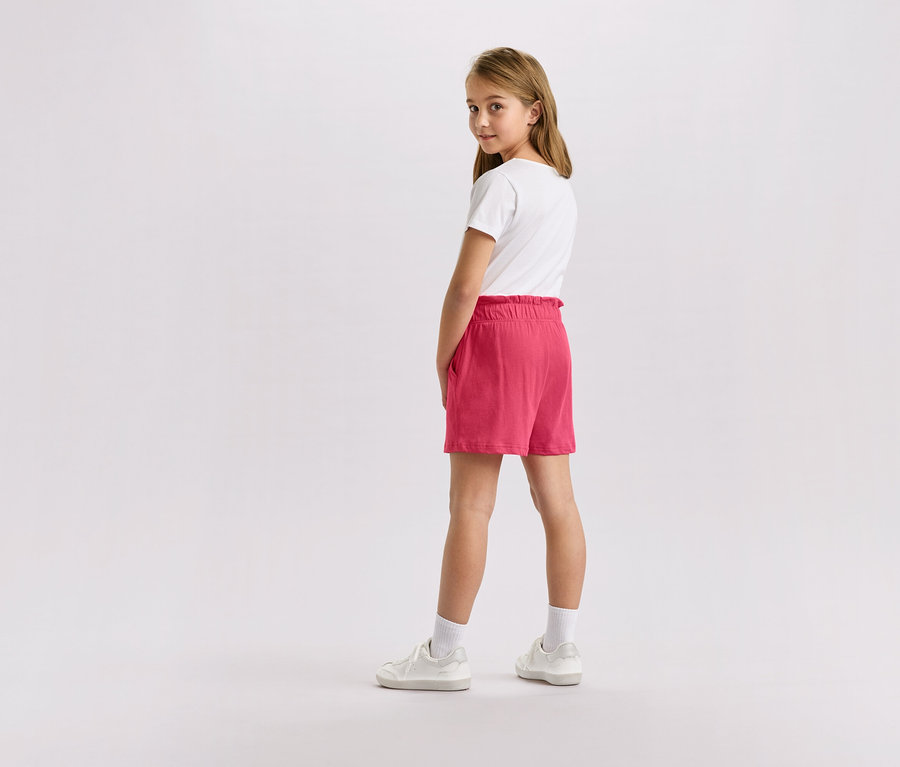 Egy szőke hajú lány fehér pólóban, rózsaszín rövidnadrágban és fehér tornacipőben, aki a kamerába néz.