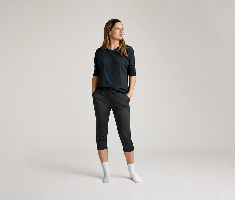 Nő fekete 3/4-es sportpólóban és sötét nadrágban, keze a zsebében, fehér zokni, világos háttér előtt.