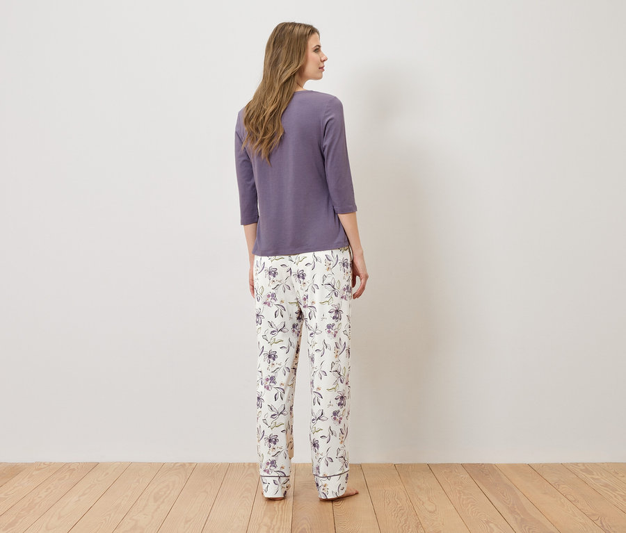 A nő háttal áll a kamerának, lila pizsamafelsőt és virágmintás fehér pizsamanadrágot visel.