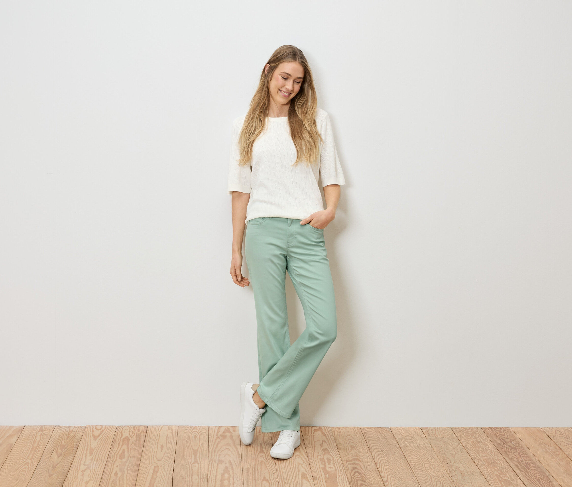 Szőke hajú nő fehér felsőben, zöld nadrágban és fehér tornacipőben áll egy fa padlón, egy fehér fal előtt.