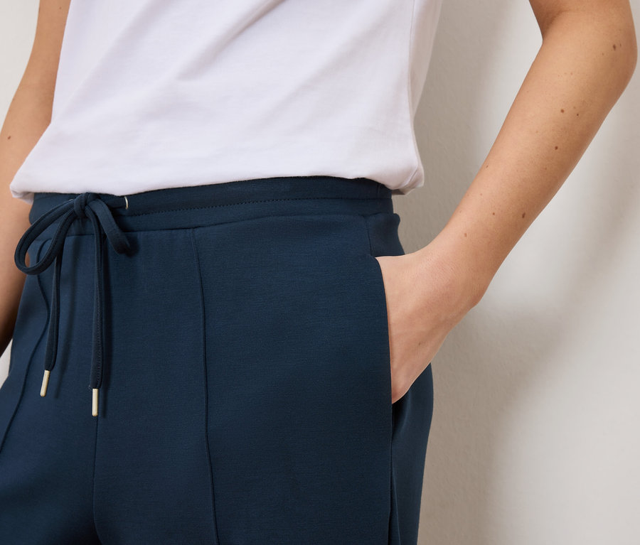 Közeli felvétel egy nőről, aki sötétkék színű, húzózsinóros, arany részletekkel ellátott melegítőnadrágot és fehér pólót visel.