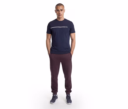 Férfi áll zsebre tett kézzel, sötétkék pólót visel fehér csíkkal és Fila logóval, bordó Fila melegítőnadrágot és sötétkék sportcipőt.
