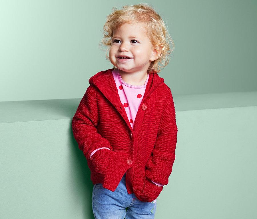 Mosolygó kisgyermek piros kötött kardigánt és farmert visel.