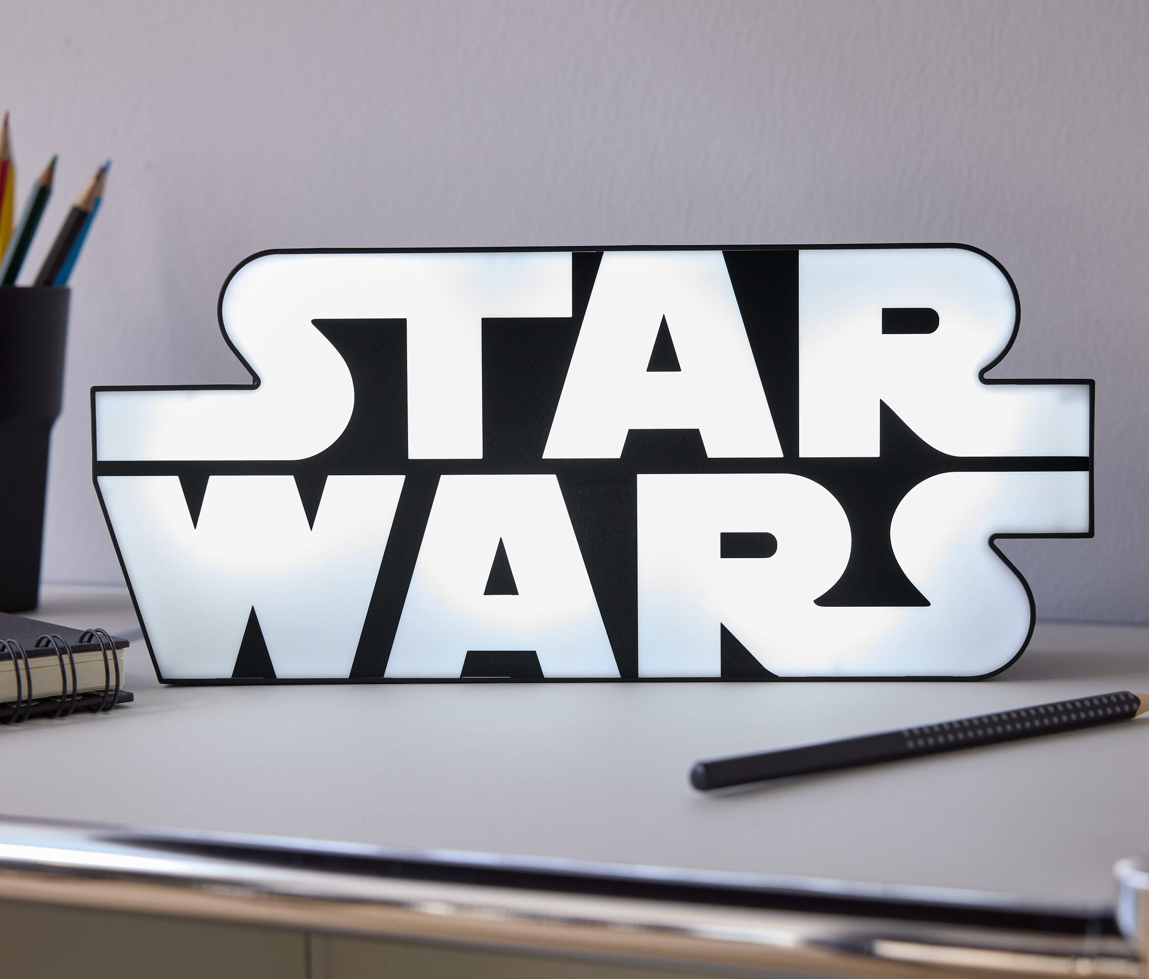 Az asztalon egy világító Star Wars felirat áll, mellette egy ceruza, egy notesz és egy pohár színes ceruza.