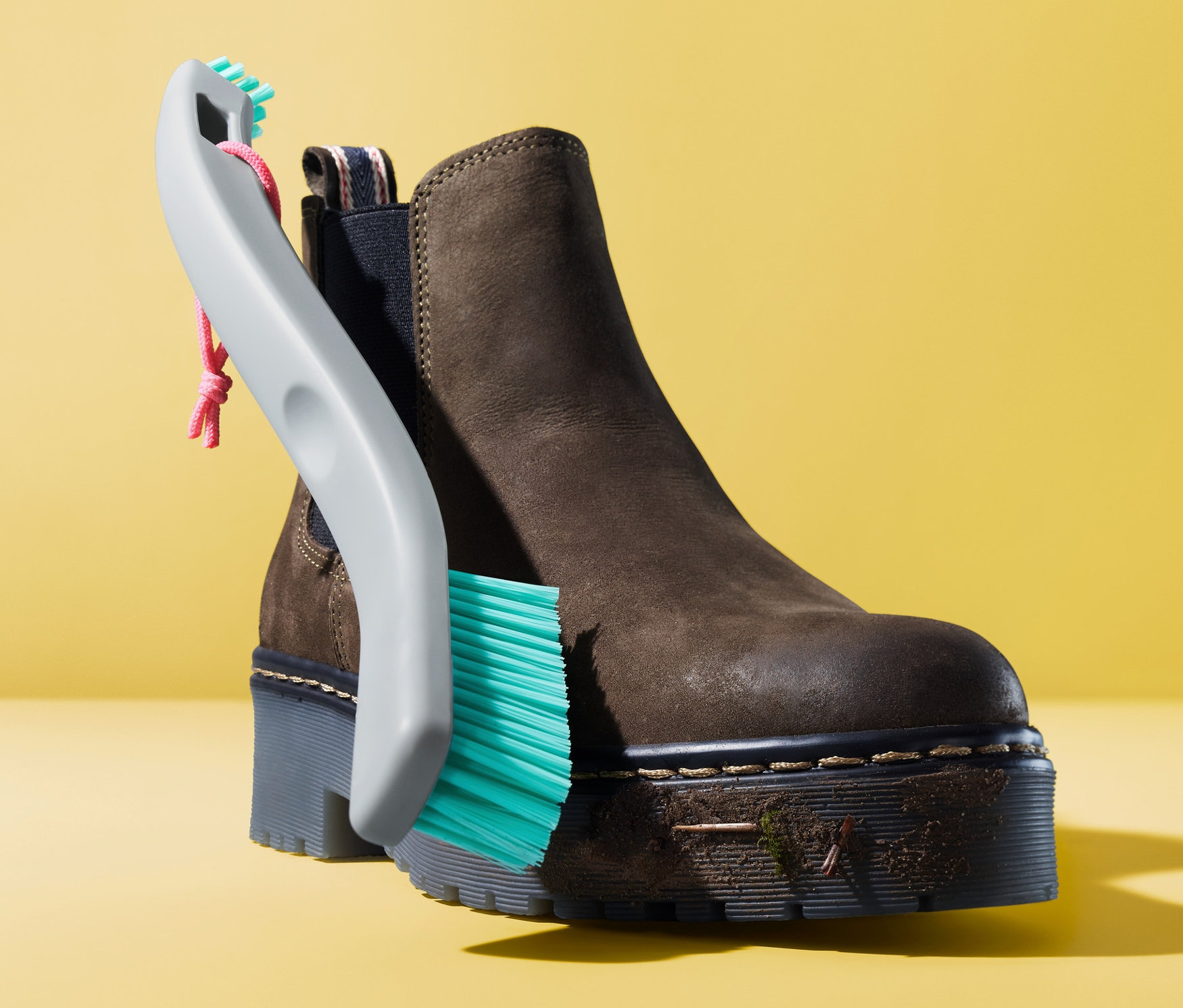 Egy barna bokacsizmát tisztítanak egy 2 az 1-ben cipőtisztító kefével sárga háttér előtt.