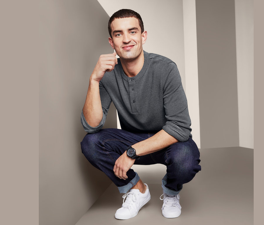 Férfi szürke, jacquard henley felsőben, farmerban és fehér cipőben pózol.