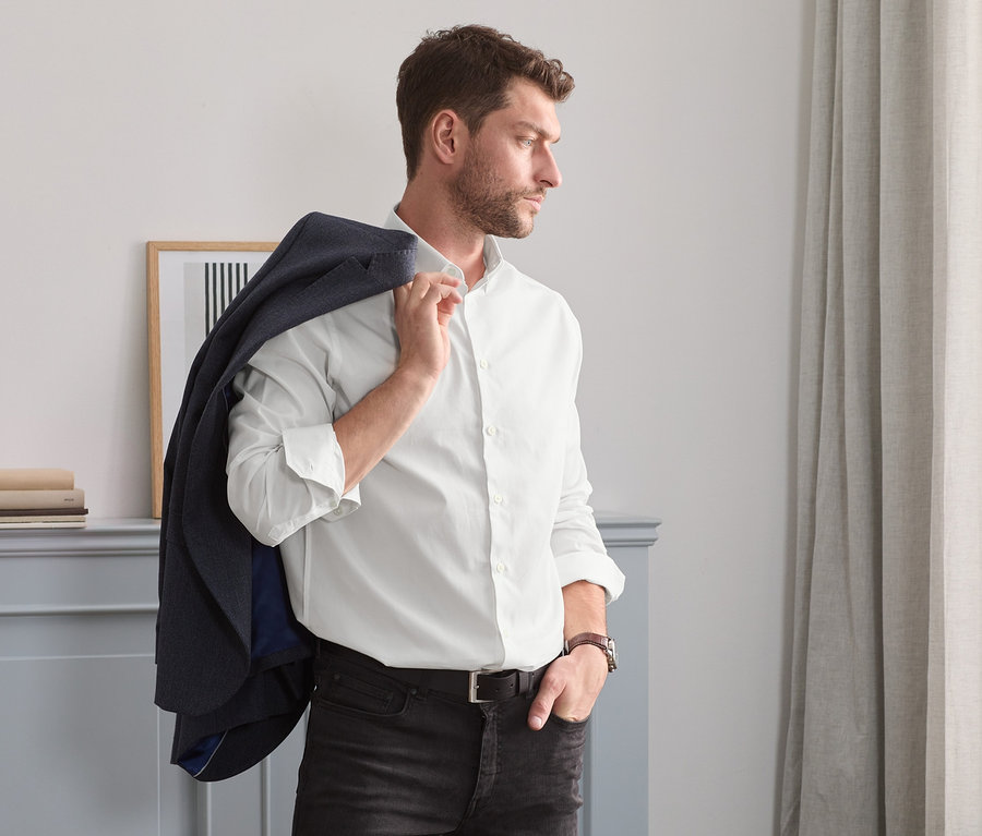 Barna hajú, borostás férfi zakót visel a vállán, és egy fehér, comfort fit oxford inget.