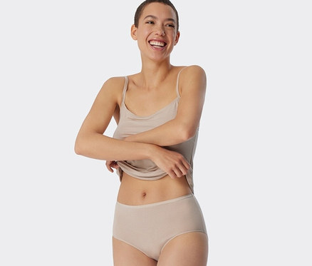 Egy mosolygó nő fogja a trikója szegélyét. Homokszínű, csíkos SCHIESSER midi alsót visel.