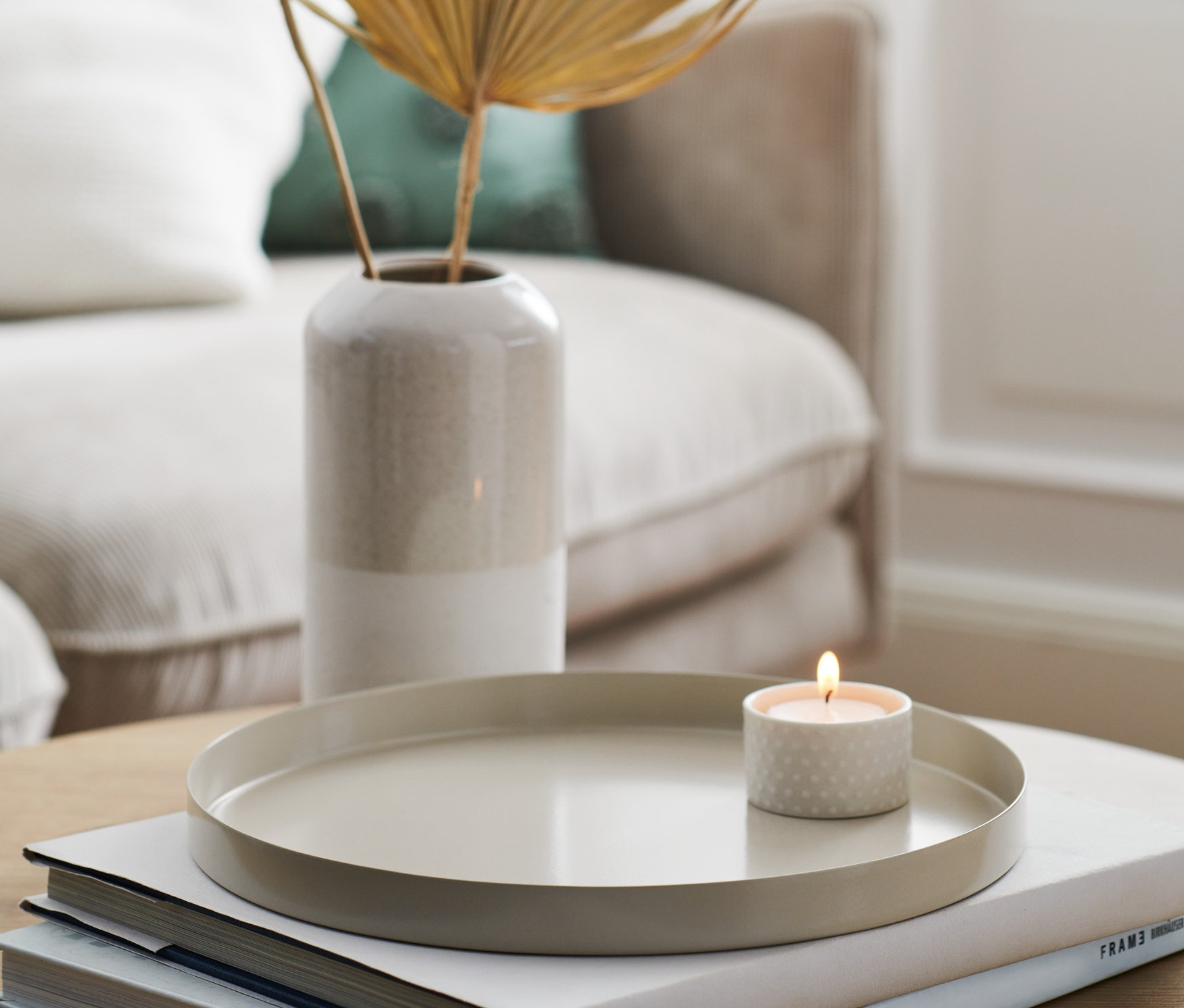 Bézs dísztálca égő gyertyával könyveken, növényekkel teli váza mellett.