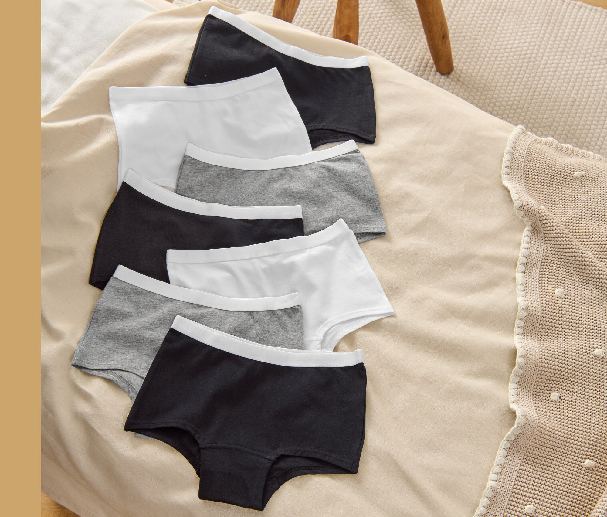 Hét gyerek alsó (7 gyerek alsó, szürke-fehér-fekete) fekszik egy bézs takarón egy fa szék mellett.