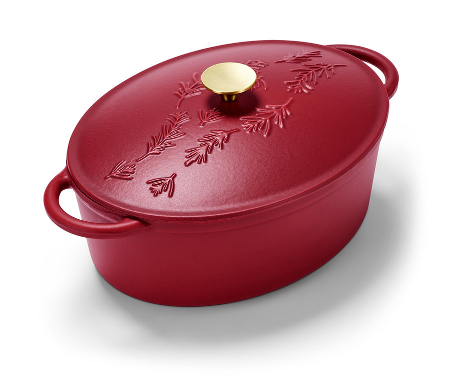 Egy piros, ovális öntöttvas sütőedény fenyőág mintával és arany fogantyúval.