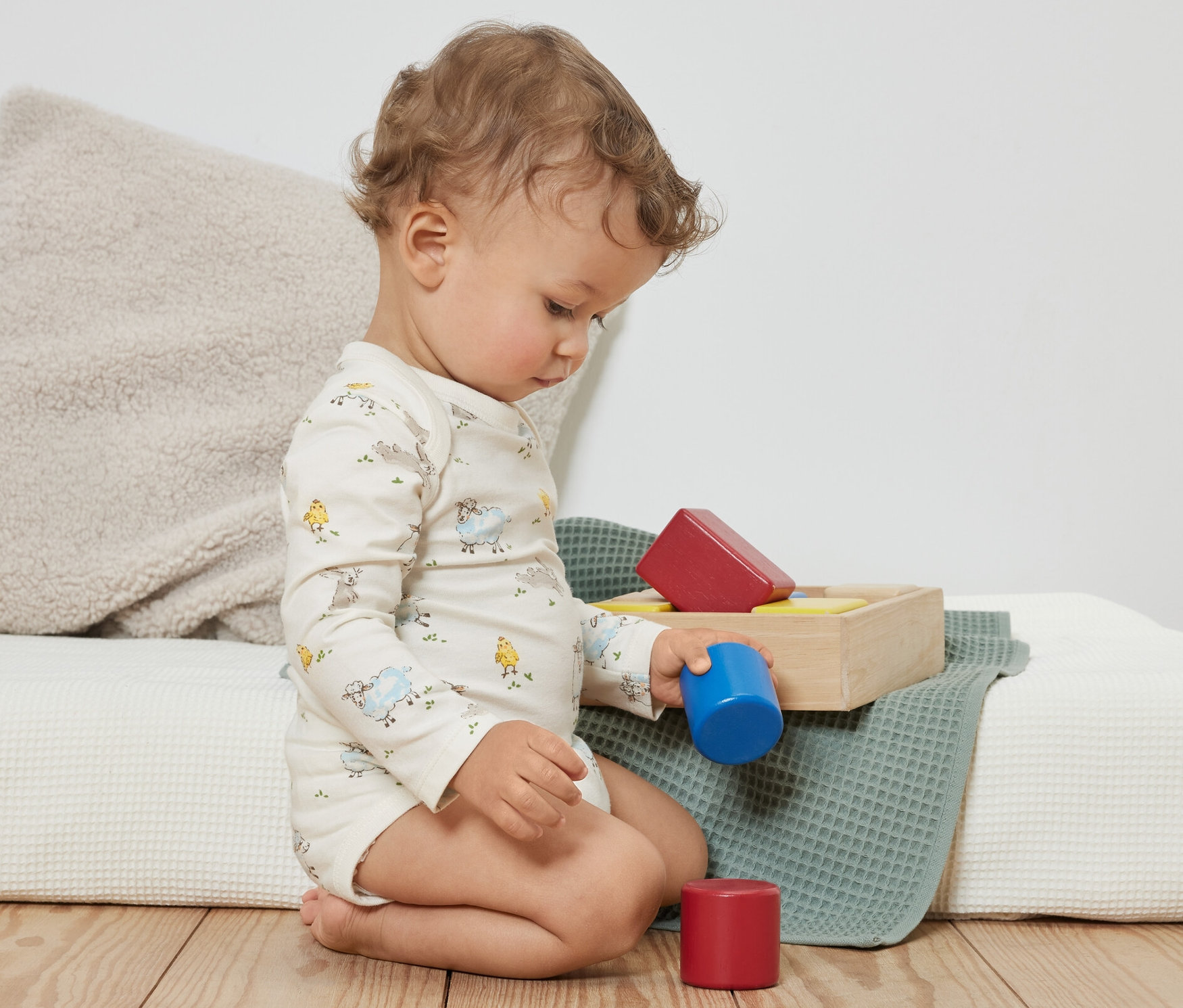 Egy göndör kisgyermek ül a padlón és színes építőkockákkal játszik. Kék színű, 3 állítható hosszúságú body csecsemőknek van rajta.
