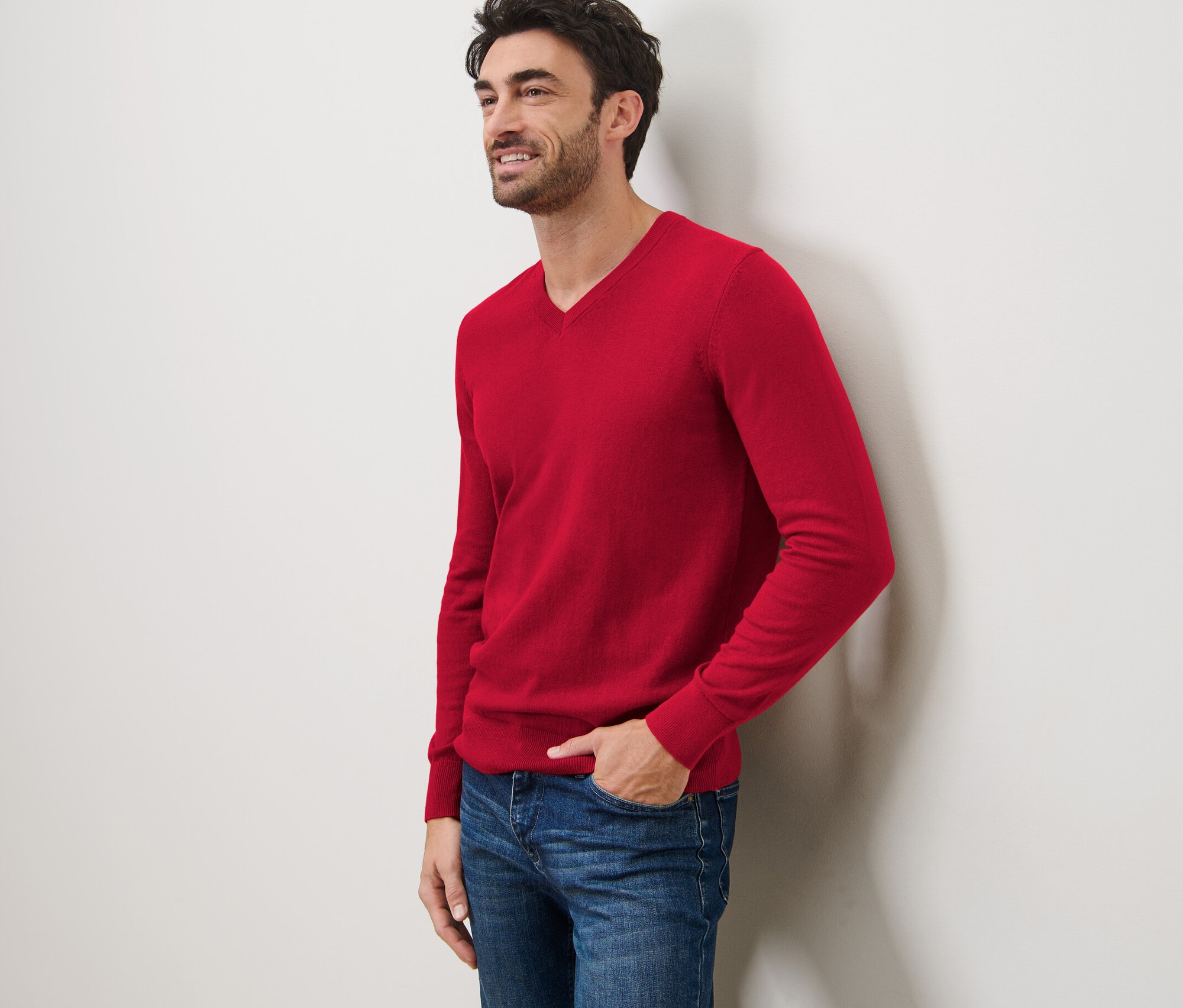 Egy mosolygó férfi a falnak dőlve piros V-nyakú pulóvert és farmert visel.