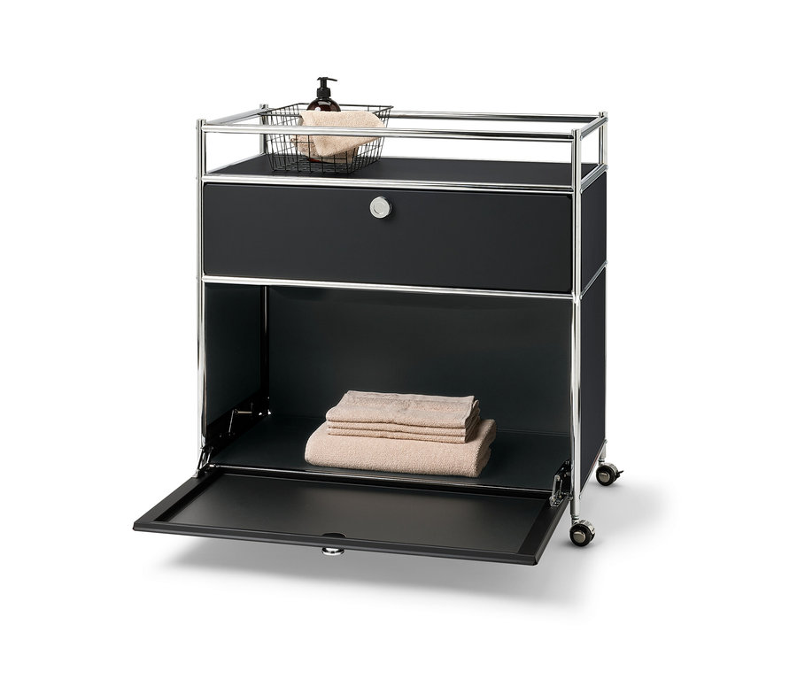 »CN3« fém komód egy fiókkal, lenyíló ajtóval és kerettel. A tetején kosár törölközővel és pumpás üveggel. Az alsó polcon összehajtott törölközők.