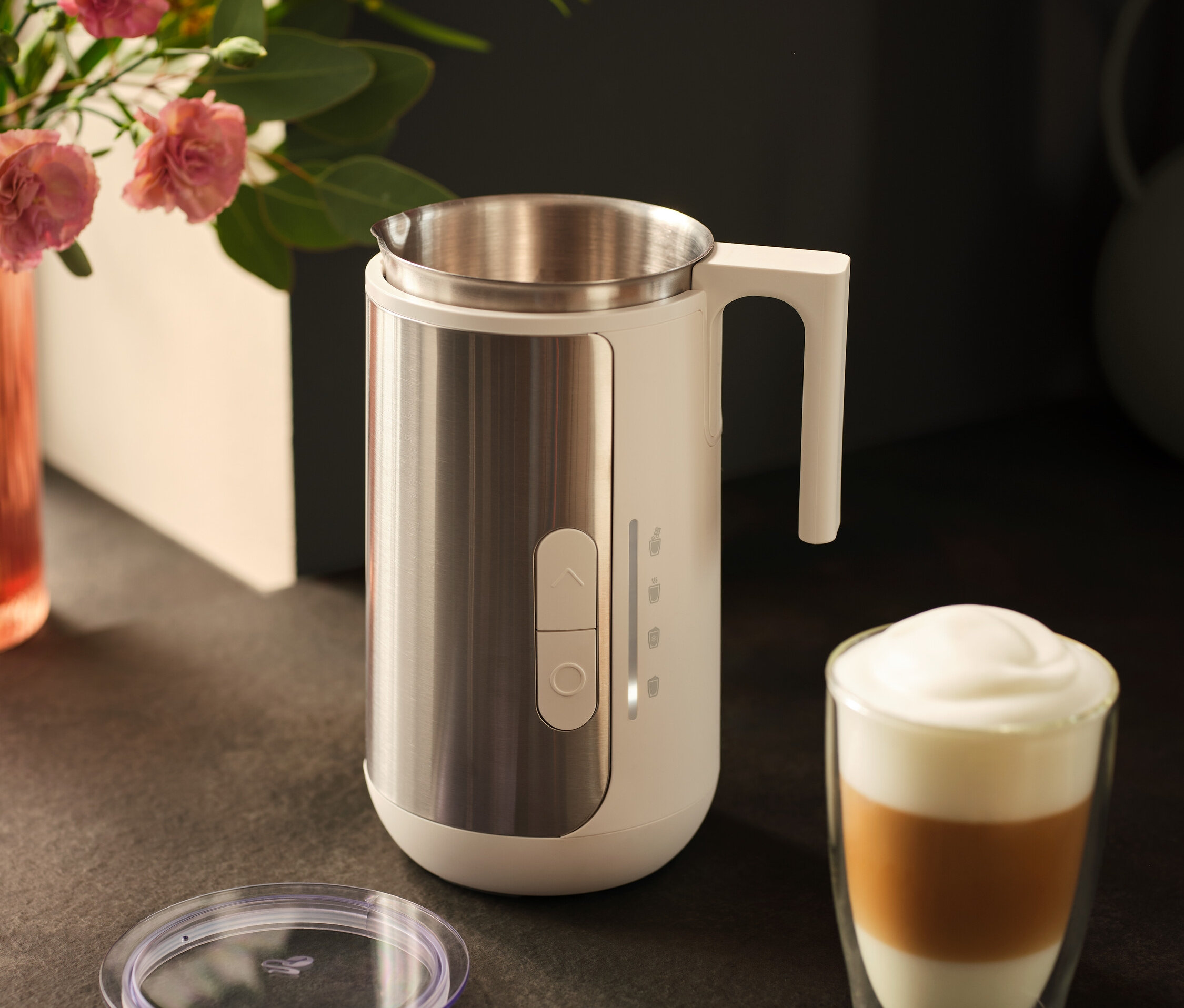 Egy rozsdamentes acélból készült tejhabosító egy latte kávéspohár és egy fedő mellett áll egy sötét pulton.