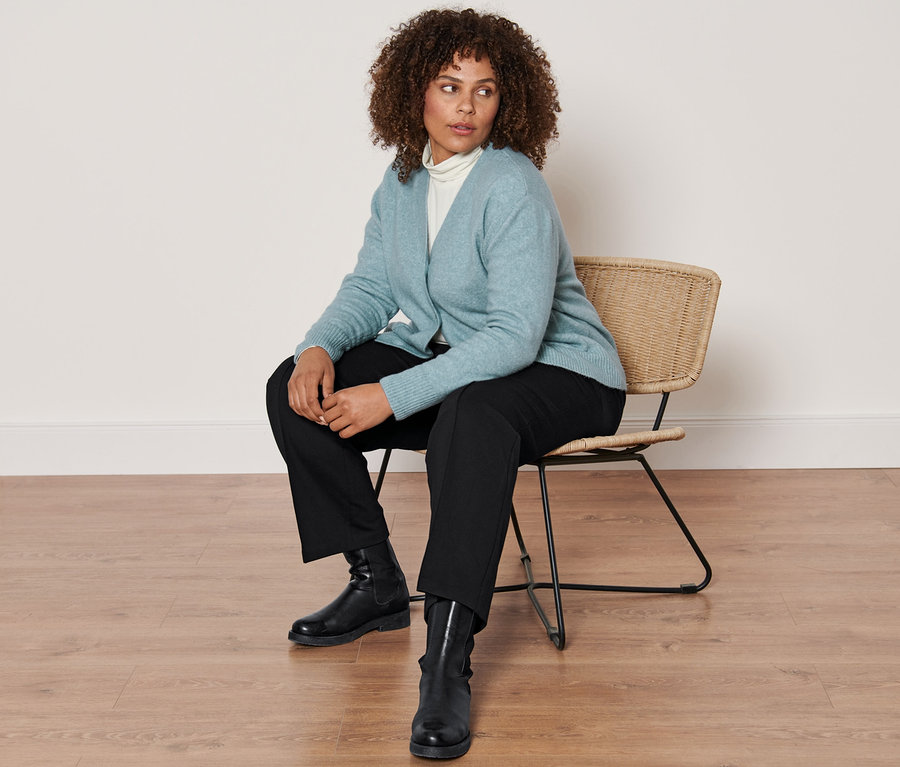 Nő ül egy széken, rajta női kötött, gyapjúval kardigán, krém színű, hosszú ujjú garbónyakú felső, fekete dzsörzénadrág levarrt hajtással és fekete női bőrcsizma.