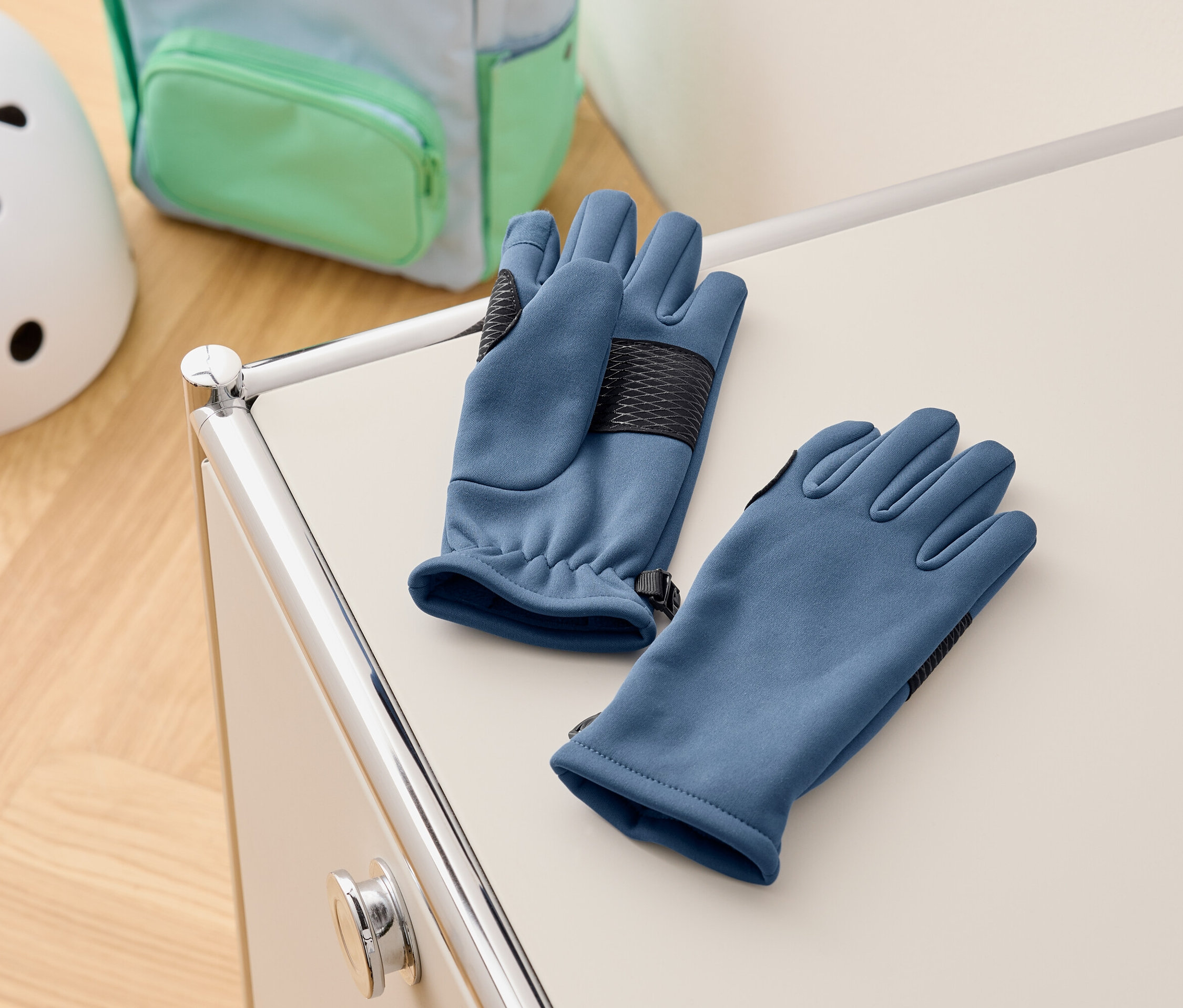 Kék kesztyűk fekszenek egy fehér szekrényen, a háttérben egy hátizsák és egy sisak.