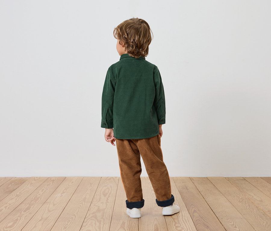 Egy fiú zöld ingben és barna nadrágban áll háttal a nézőnek egy fapadlón.