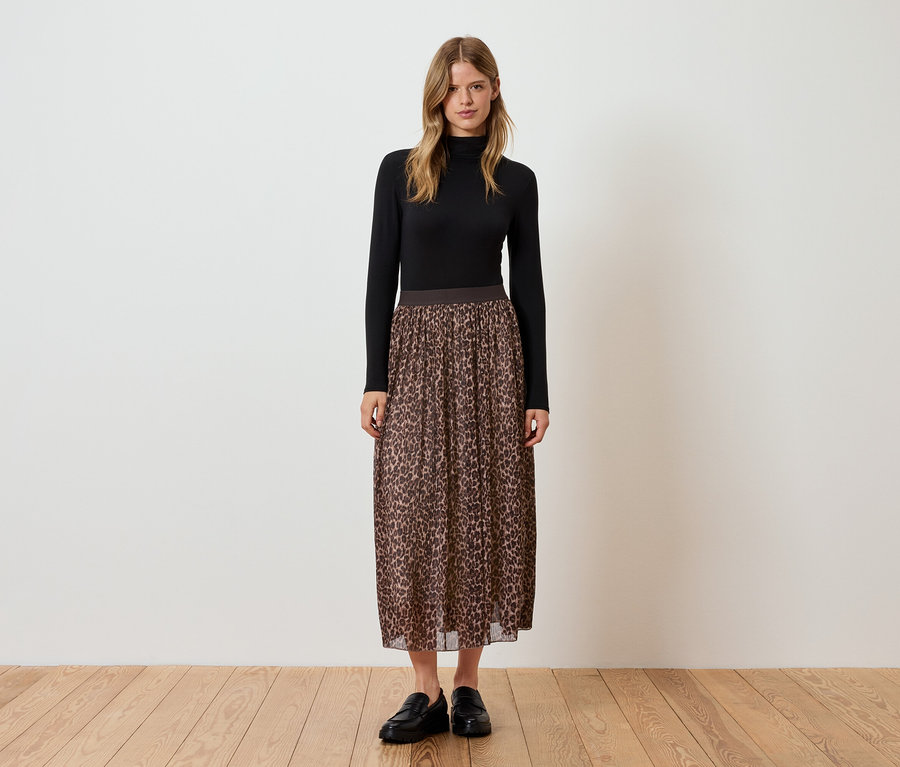 Egy nő áll fekete garbóban, barna, állatmintás rakott szoknyában és fekete cipőben.