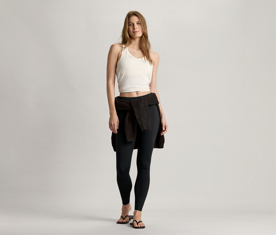 Nő áll, és fehér, varrásmentes sportmelltartót, fekete leggingset és a derekára kötött kardigánt visel.