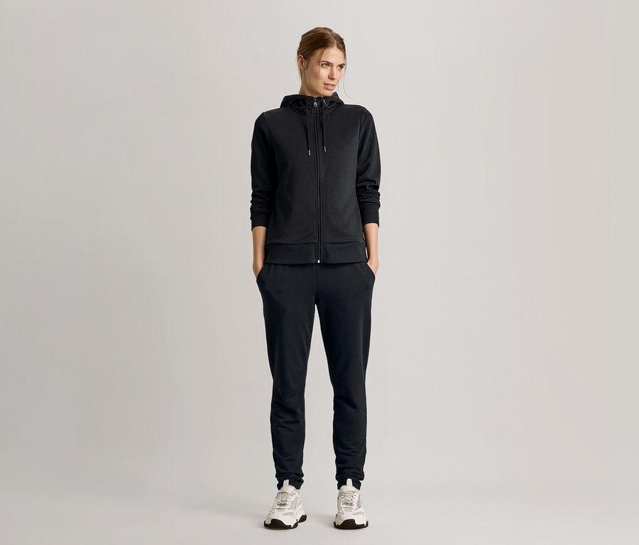 Nő áll kezével a zsebében, fekete kapucnis pulóveres tréningruhát és tornacipőt visel.