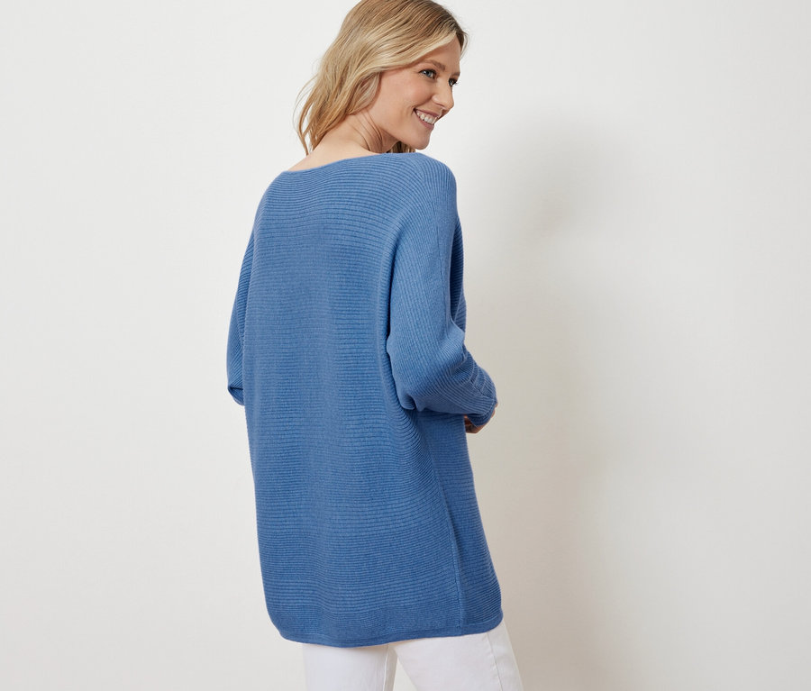 Szőke nő mosolyog a válla fölött, kék, finom kötésű pulóvert és fehér nadrágot visel.