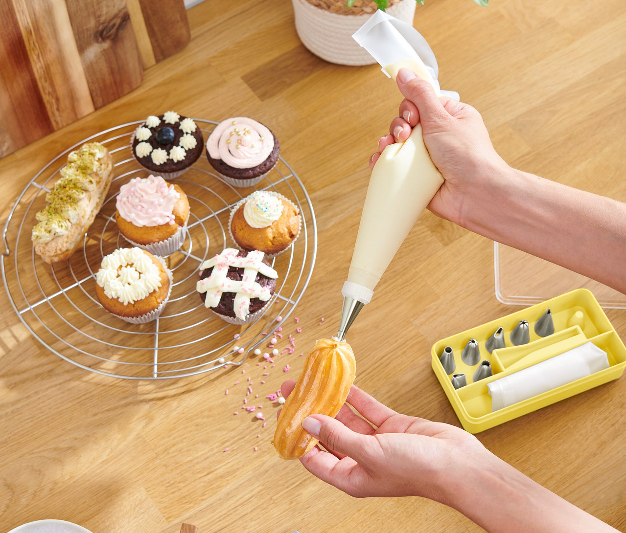 Egy személy egy tésztát díszít habzsákkal, muffinok és egy sárga doboz a csövekkel egy fa asztalon fekszenek.