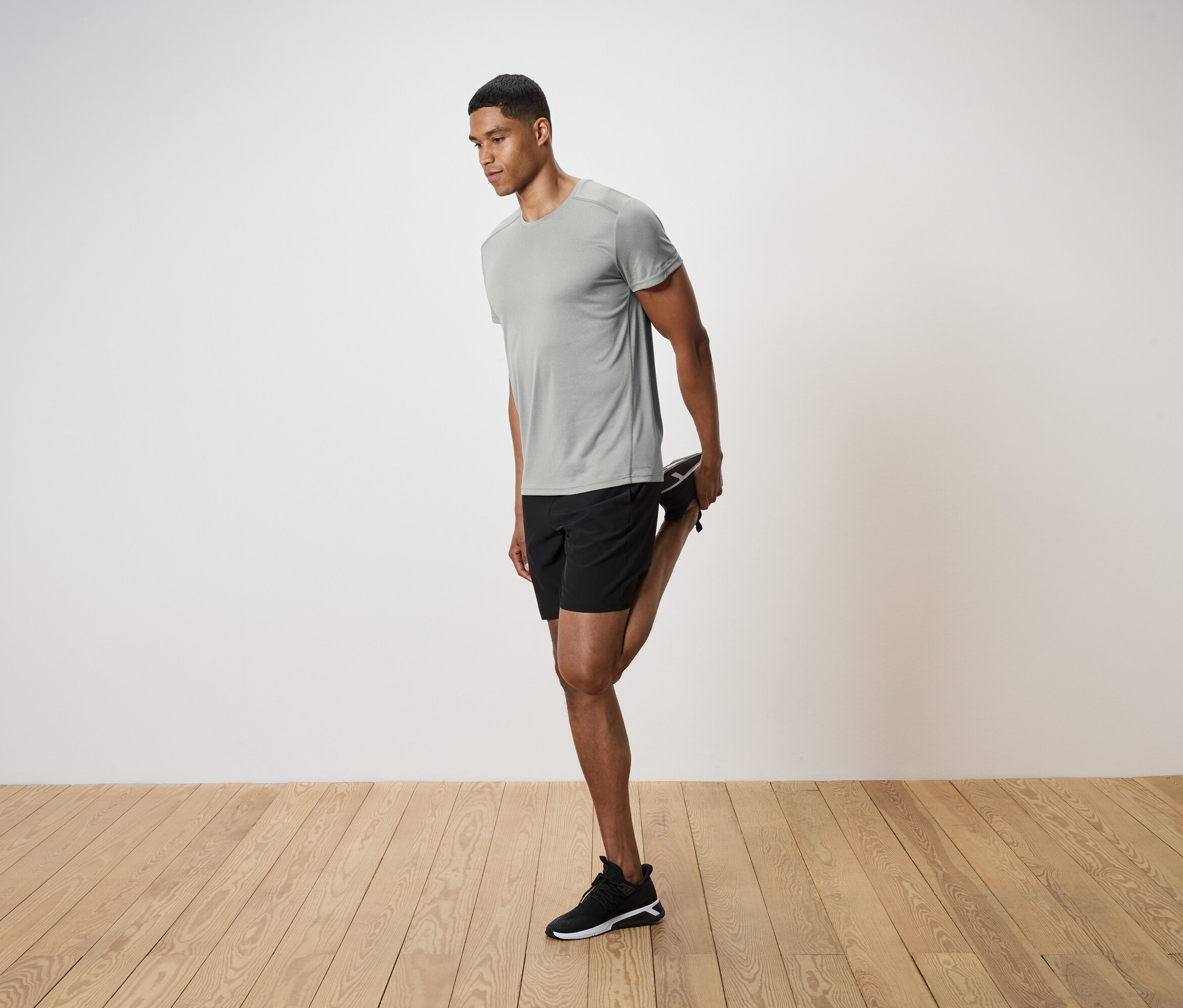 Férfi szürke pólóban és fekete rövidnadrágban nyújtja a lábát egy fapadlón.