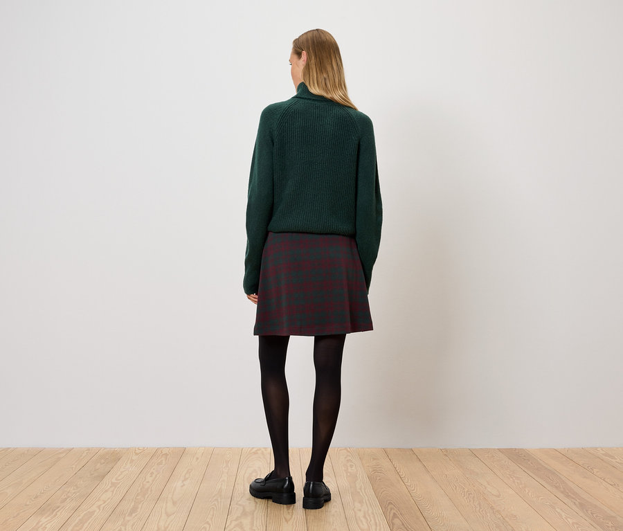 Egy nő háttal áll a kamerának, zöld pulóvert, kockás szoknyát és fekete harisnyát visel.
