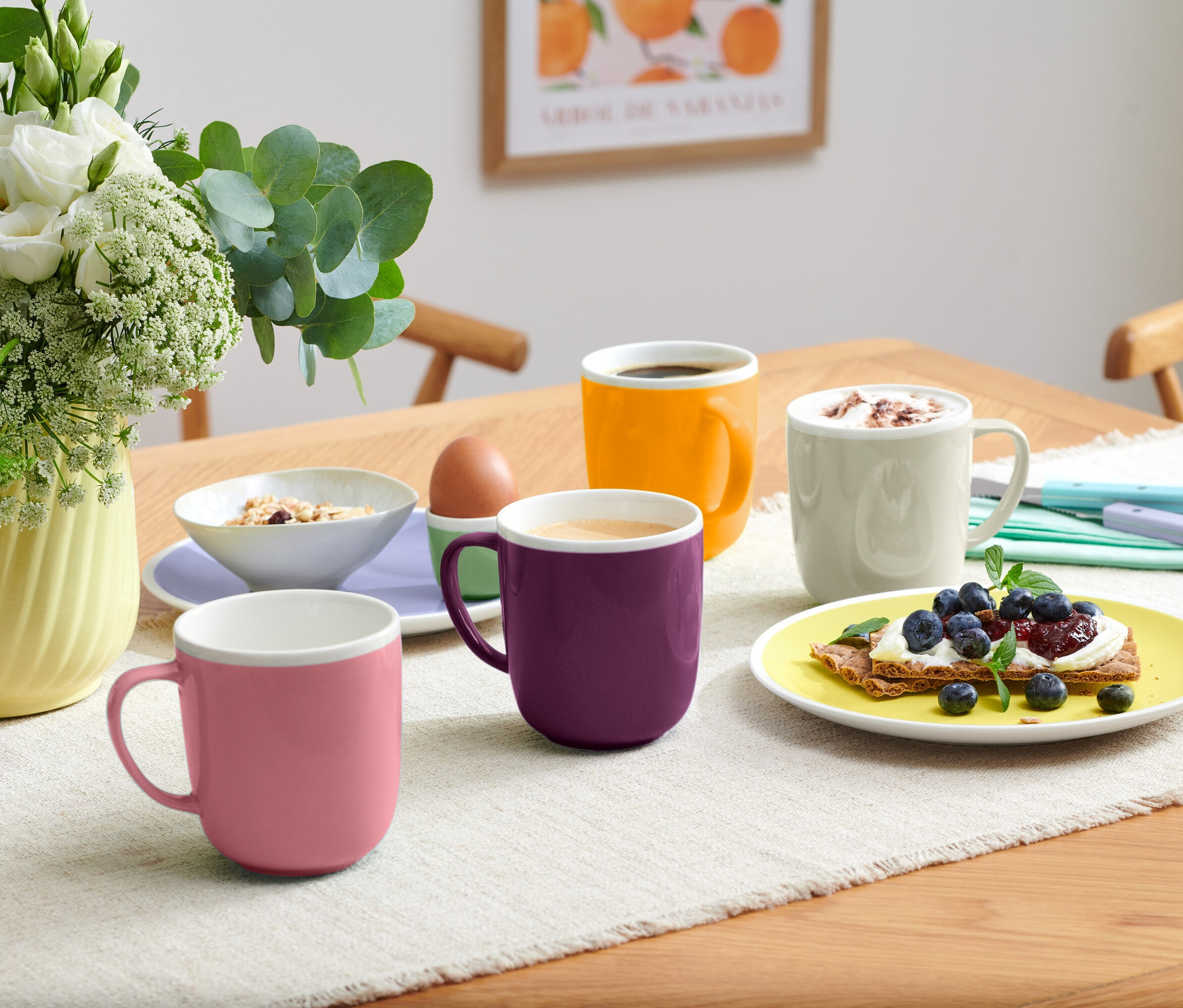 Az asztalon négy különböző színű bögre, egy müzlis tál, egy tojás egy tojástartóban és egy tányér áfonyás pirítós.