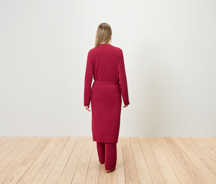 Hátulnézet egy szőke hajú nőről, aki bordó köntöst és nadrágot visel egy fapadlón.