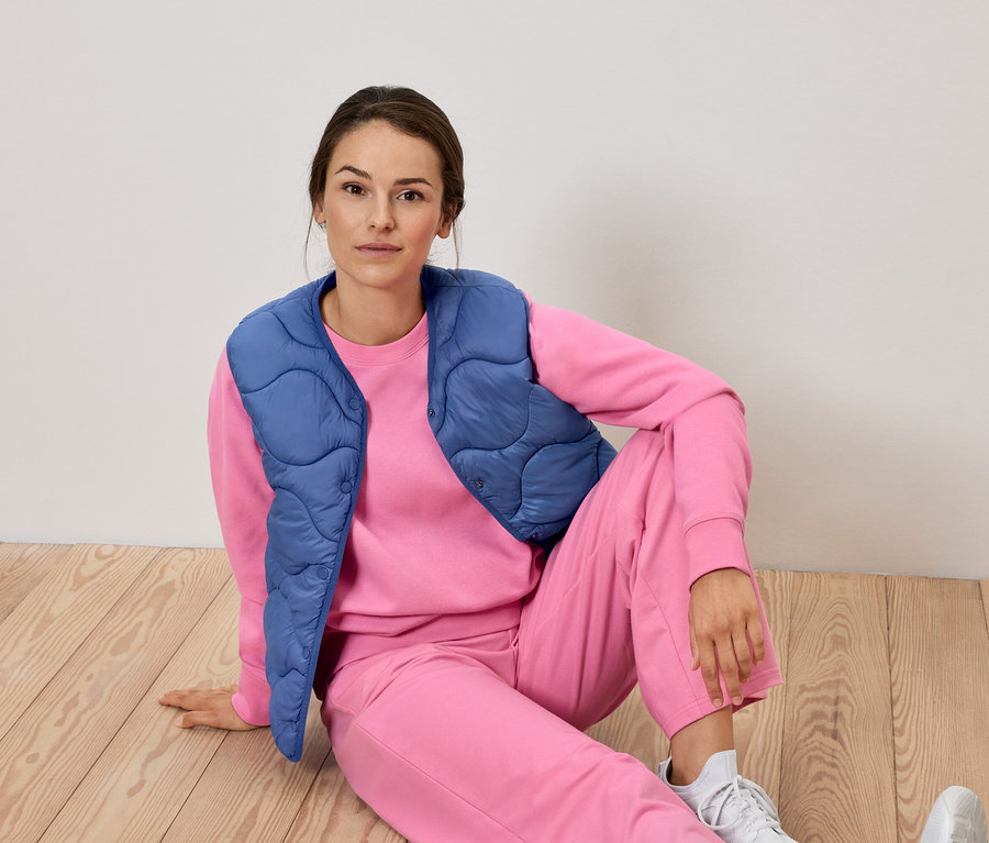 Nő ül rózsaszín tréningruhában és kék mellényben a padlón.