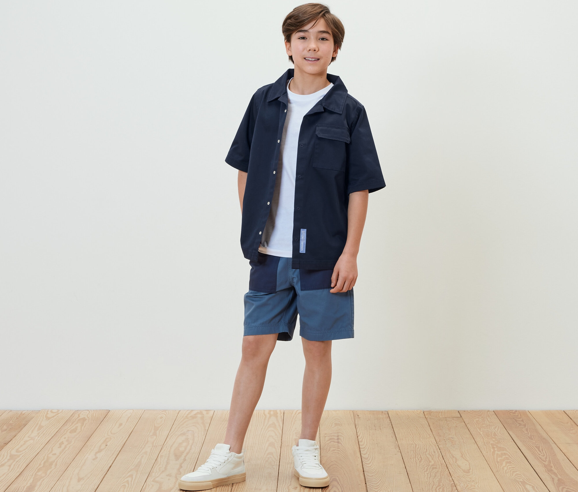 Egy fiú áll, és egy lezser gyerekinget, rövidnadrágot és tornacipőt visel.