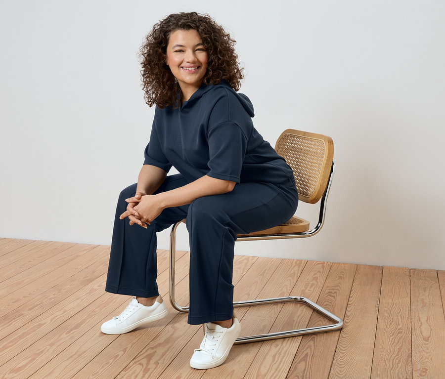 Göndör hajú nő ül egy széken, sötétkék tréningruhát és fehér tornacipőt visel.