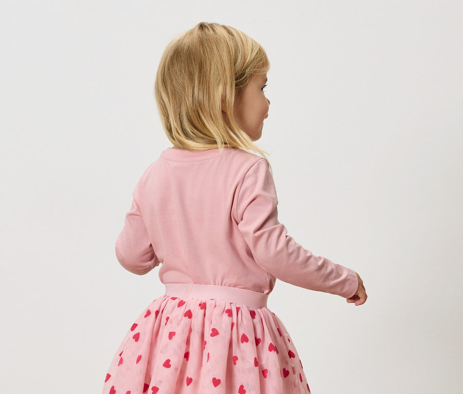 Egy kis szőke lány rózsaszín, hosszú ujjú pólót és rózsaszín tüllszoknyát visel piros szívekkel.