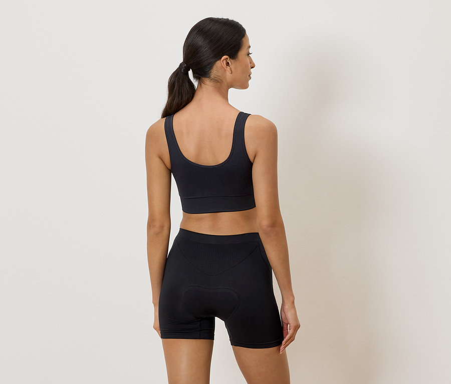 Hátulról látható nő fekete sportmelltartóban és biciklisnadrágban, semleges háttér előtt.