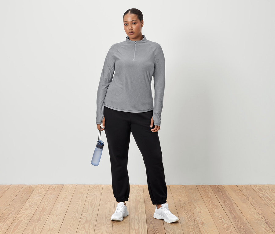 Egy nő áll egy fehér fal előtt, és egy kék vizespalackot tart a kezében. Szürke, hosszú ujjú pólót, fekete jogging nadrágot és fehér tornacipőt visel.