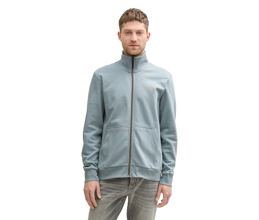 A férfi szürkés menta színű Tom Tailor állógalléros szabadidőfelsőt és szürke farmert visel.