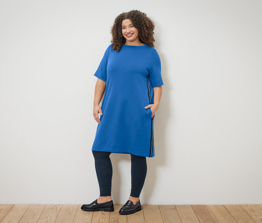 Göndör hajú nő cipzáras kék ruhában és leggingsben pózol a fal előtt.